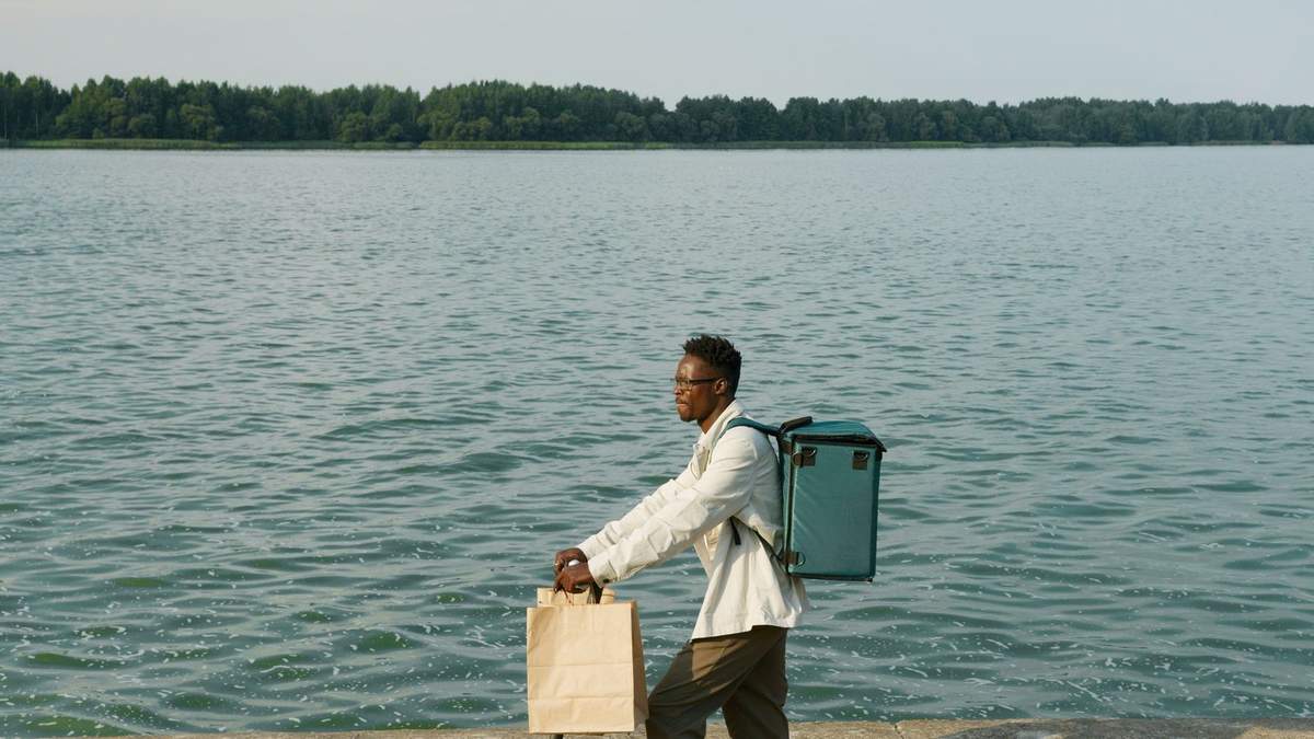 Homme noir marchant avec deux sacs près d'un lac, arbres en arrière-plan Homme noir marchant avec deux sacs près d'un lac, arbres en arrière-plan