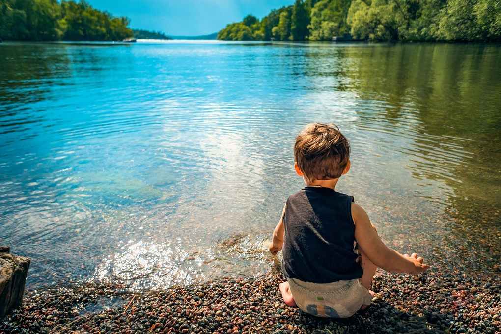 Enfant en noir et blanc assis sur des galets près d'un lac, les bras tendus vers l'eau Enfant en noir et blanc assis sur des galets près d'un lac, les bras tendus vers l'eau